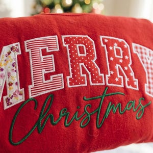 Op de afbeelding: Een rood sweatshirt met het woord MERRY in grote, van stof gemaakte applicatie-blokletters met diverse patronen zoals bloemen, strepen, stippen en sterren, met het woord Christmas eronder geborduurd in groen cursief schrift.