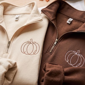 May include: Two quarter-zip sweatshirts, one in cream and one in dark brown. The cream sweatshirt features a brown embroidered pumpkin outline, while the dark brown sweatshirt features a white embroidered pumpkin outline on the chest area.