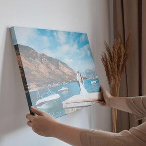 Pu&ograve; includere: Stampa su tela quadrata raffigurante un paesaggio acquatico con barche, montagne e una coppia su un molo. La stampa &egrave; presentata contro un muro bianco. La tela misura circa 40 cm quadrati, mostrando i colori e i dettagli.