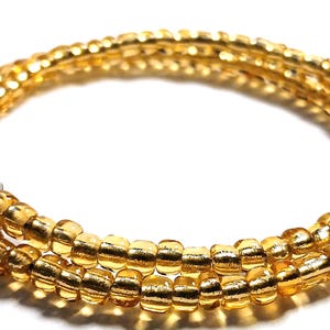 Puede incluir: Una pulsera enrollada, de cuentas amarillo dorado. Las cuentas, peque&ntilde;as y cil&iacute;ndricas, est&aacute;n ensartadas para formar m&uacute;ltiples hebras. La pulsera tiene un cierre met&aacute;lico. El fondo es blanco.