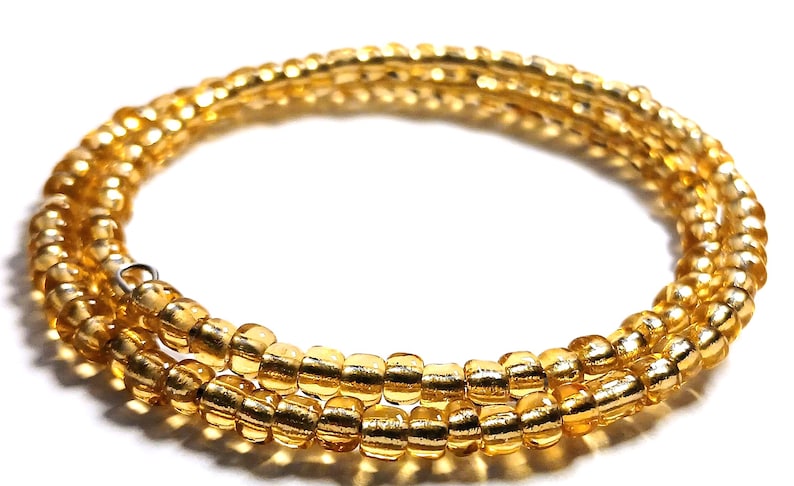 Puede incluir: Una pulsera enrollada, de cuentas amarillo dorado. Las cuentas, peque&ntilde;as y cil&iacute;ndricas, est&aacute;n ensartadas para formar m&uacute;ltiples hebras. La pulsera tiene un cierre met&aacute;lico. El fondo es blanco.