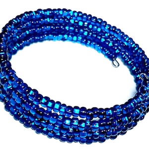 May include: A coiled bracelet made of small, round, deep blue beads. The bracelet wraps around multiple times, creating a layered effect. The beads have a glossy finish, reflecting light. The clasp is a small silver-colored metal hook.
