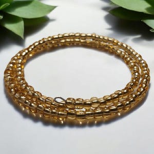 Puede incluir: Un brazalete enrollado de cuentas doradas. Las cuentas, peque&ntilde;as y redondas, est&aacute;n ensartadas para formar m&uacute;ltiples hebras. El brazalete se exhibe sobre una superficie clara, con follaje verde en el fondo.