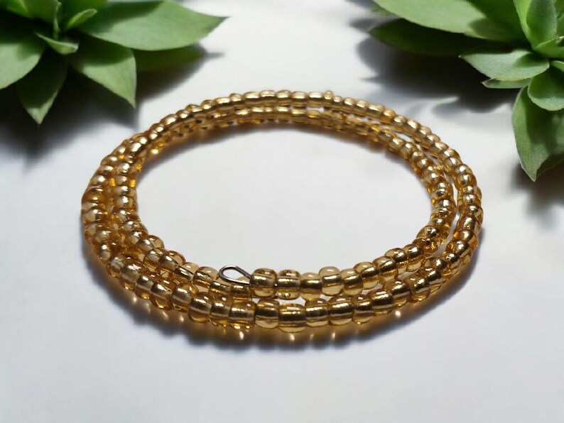 Puede incluir: Un brazalete enrollado de cuentas doradas. Las cuentas, peque&ntilde;as y redondas, est&aacute;n ensartadas para formar m&uacute;ltiples hebras. El brazalete se exhibe sobre una superficie clara, con follaje verde en el fondo.