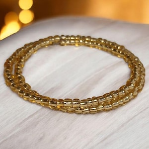 Puede incluir: Una pulsera de cuentas doradas, enrollada en m&uacute;ltiples hebras. Las cuentas son peque&ntilde;as y de tama&ntilde;o uniforme, creando un dise&ntilde;o circular texturizado. La pulsera se muestra sobre una superficie blanca, con iluminaci&oacute;n suave.