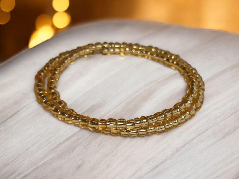 Puede incluir: Una pulsera de cuentas doradas, enrollada en m&uacute;ltiples hebras. Las cuentas son peque&ntilde;as y de tama&ntilde;o uniforme, creando un dise&ntilde;o circular texturizado. La pulsera se muestra sobre una superficie blanca, con iluminaci&oacute;n suave.