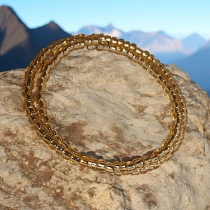 Puede incluir: Un brazalete dorado de cuentas, enrollado en m&uacute;ltiples hebras, descansa sobre una roca texturizada. Las cuentas tienen un tono &aacute;mbar c&aacute;lido. El fondo presenta una cordillera bajo un cielo azul claro.