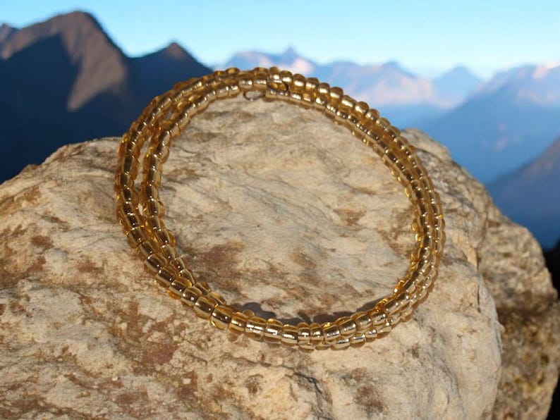 Puede incluir: Un brazalete dorado de cuentas, enrollado en m&uacute;ltiples hebras, descansa sobre una roca texturizada. Las cuentas tienen un tono &aacute;mbar c&aacute;lido. El fondo presenta una cordillera bajo un cielo azul claro.