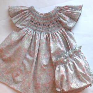 Könnte beinhalten: Ein zweiteiliges Baby-Set aus Kleid und Bloomers mit hellrosa und grünem Blumenmuster. Das Kleid hat ein smockverziertes Oberteil, flatternde Ärmel und einen runden Ausschnitt. Die Bloomers haben eine Rüschenborte. Der Stoff hat ein zartes Blumenmuster.