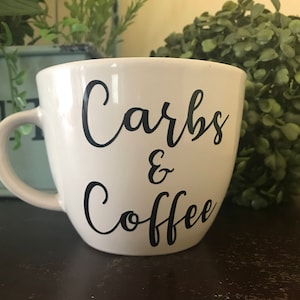 May include: White ceramic coffee mug with black lettering that reads "Carbs & Coffee".