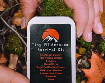 May include: A small, silver tin with a red and black logo that says "Tiny Wilderness Survival Kit". The tin contains a list of survival items, including an emergency blanket, a high volume whistle, a compass, a signal mirror, water purification powder, waterproof matches, antibacterial cream, bandaids, and twine.