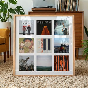 May include: A framed collection of nine album covers, each with unique artwork and titles. The frame is light wood, and the album covers feature various colors and designs, including text and illustrations. A record player and vinyl records are visible in the background.