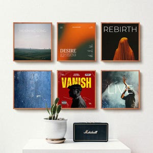 Può includere: Sei stampe incorniciate di copertine di album con vari disegni e testo, tra cui "Morning Song", "Desire" e "Rebirth". Sono presenti anche un piccolo cactus in un vaso bianco e un altoparlante Marshall nero.