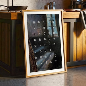 May include: A framed art print with a light wood frame. The artwork features a dark, impressionistic painting with the words "NOTES - FROM UNDER GROUND - FYO ODOR - DOSTO EVSKY" in white text. The print is leaning against a wooden cabinet.