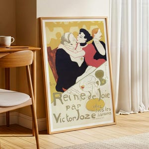 May include: A framed vintage poster with a yellow background and a scene of three people. The poster has the words "Reine de Joie" and "Victor Joze" in green and gold lettering. The frame is made of light brown wood.