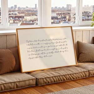 May include: A framed print with a light beige background and handwritten black text. The frame is a warm brown colour. The print rests on a brown cushion, with a city view visible in the background.