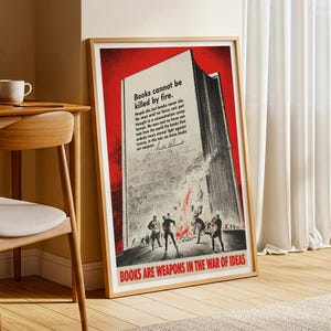 May include: A framed vintage poster with a red and white design. The poster depicts a burning book with figures in the foreground. Text includes "Books cannot be killed by fire" and "Books are weapons in the war of ideas."