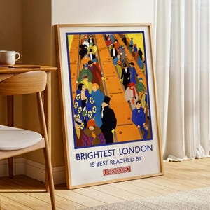 Op de afbeelding: Ingelijste vintage poster met de tekst "Brightest London is Best Reached By Underground". De poster toont een kleurrijke illustratie van mensen op een perron, met een gele en oranje achtergrond en een blauwe rand. De poster zit in een licht houten frame.