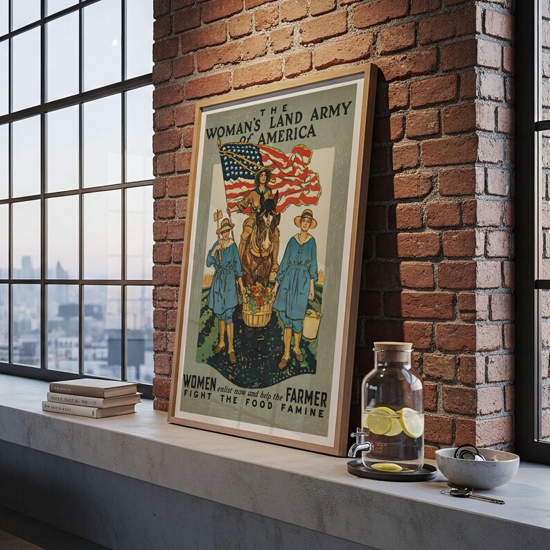 May include: A framed vintage poster with a wooden frame. The poster features an illustration of women in blue dresses and hats, with an American flag and text that reads "The Woman's Land Army of America". The poster is leaning against a brick wall.