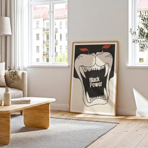 May include: A framed poster with a black panther illustration. The panther's mouth is open, revealing sharp white teeth. The text "Black Power" is in the center. The poster has a gold frame and is leaning against a white wall.