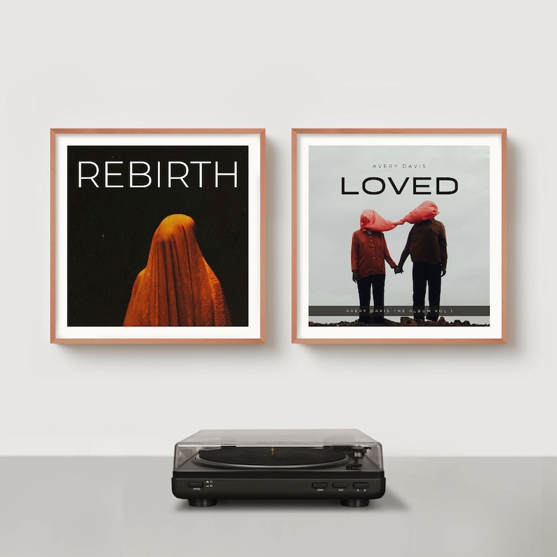 May include: Two framed album art prints with rose gold frames hang above a black turntable. The left print says "REBIRTH" with an orange figure on a black background. The right print says "LOVED" with two figures holding hands.