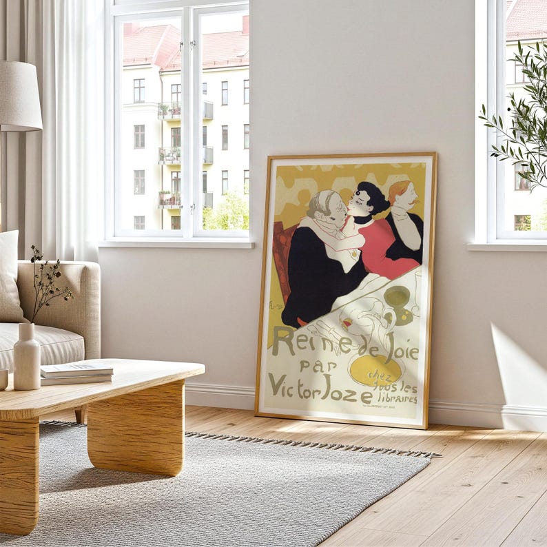 May include: A framed vintage poster titled "Reine de Joie" by Victor Joze. The poster features a colorful illustration of three figures in a cafe setting. The poster is displayed in a living room with a wooden floor and a gray rug.