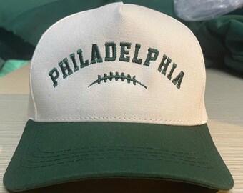 May include: A baseball cap with a white crown and a green bill. The word "PHILADELPHIA" is embroidered in green across the front, above a football graphic. The hat is made of a durable fabric.