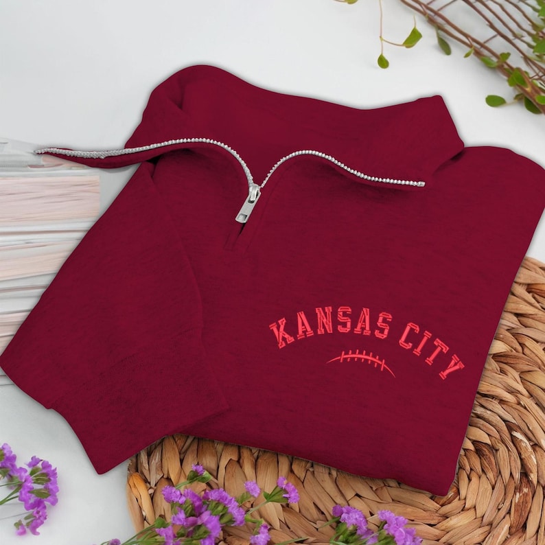 May include: A maroon quarter-zip sweatshirt with the words "KANSAS CITY" embroidered in red. The sweatshirt has a silver zipper and is folded on a woven basket.