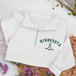 Könnte beinhalten: Weißes Sweatshirt mit Viertelreißverschluss, dem Wort "MINNESOTA" in grünen Blockbuchstaben und einer grünen Hockeyschläger-Grafik. Das Sweatshirt hat einen silbernen Reißverschluss und lange Ärmel mit gerippten Bündchen.