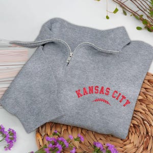 May include: A gray sweatshirt with a quarter-zip neckline. The red embroidered text "KANSAS CITY" is arched above a football graphic. The sweatshirt is folded on a woven mat with purple flowers.
