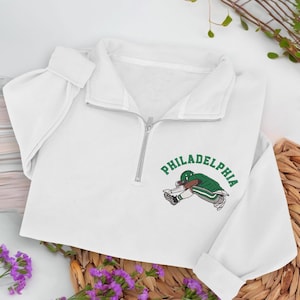 Può includere: Felpa bianca con zip a un quarto di zip argentata. La felpa presenta una grafica verde e bianca di un giocatore di football americano e la scritta "PHILADELPHIA" in verde. Le maniche sono lunghe e il materiale sembra morbido.