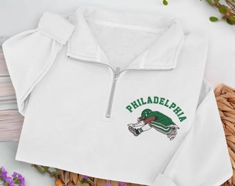 May include: White quarter-zip sweatshirt with a silver zipper. The sweatshirt features a green and white graphic of a football player and the word "PHILADELPHIA" in green. The sleeves are long and the material appears soft.