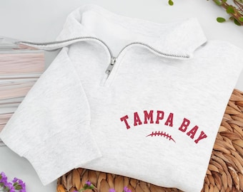 Pode incluir: Um moletom cinza claro com gola com zíper. As palavras "TAMPA BAY" são bordadas em vermelho, com um gráfico de futebol americano embaixo. O moletom está dobrado sobre uma superfície tecida, com flores roxas em primeiro plano.