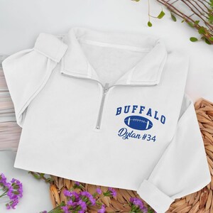 Könnte beinhalten: Weißes Sweatshirt mit Viertelreißverschluss und silbernem Reißverschluss. Das Sweatshirt zeigt das Wort "BUFFALO" über einer blauen Football-Grafik mit "Dylan #34" darunter. Die Ärmel sind umgeschlagen.