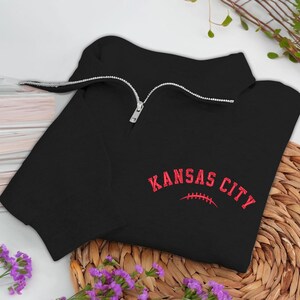 May include: Black quarter-zip sweatshirt with a silver zipper. The sweatshirt features the words "KANSAS CITY" in red, arched above a red football graphic. The garment is displayed on a woven surface with purple flowers.