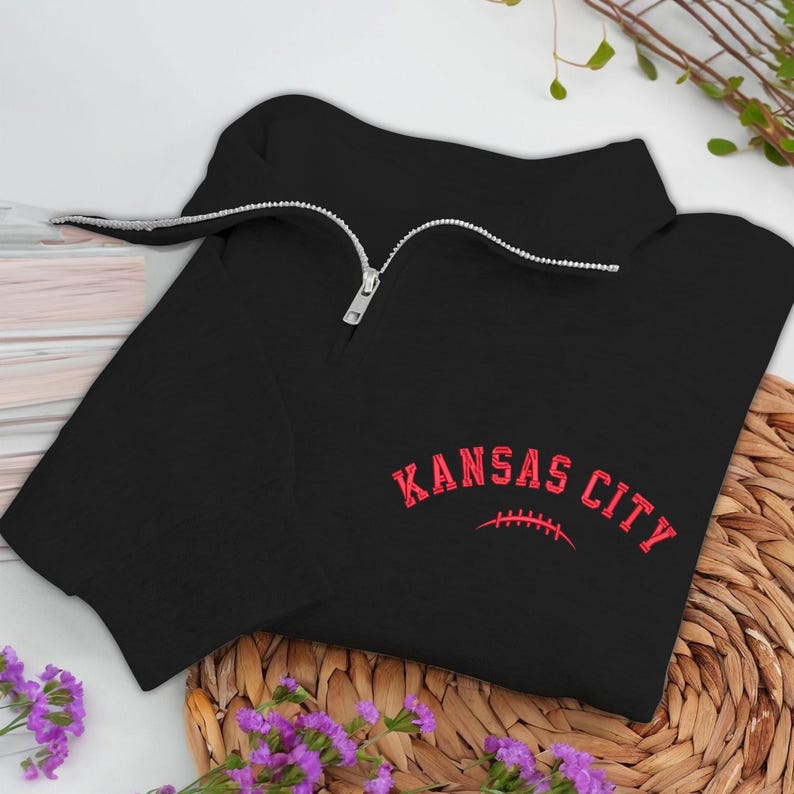 May include: Black quarter-zip sweatshirt with a silver zipper. The sweatshirt features the words "KANSAS CITY" in red, arched above a red football graphic. The garment is displayed on a woven surface with purple flowers.