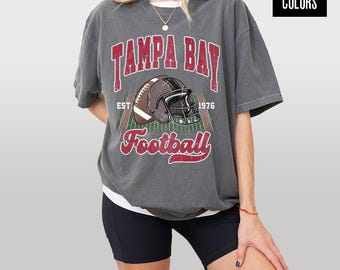 Pode incluir: Uma camiseta cinza com o texto "TAMPA BAY Football" em vermelho e branco. A camiseta apresenta um gráfico de capacete de futebol e o texto "EST. 1976". A camiseta faz parte da coleção Comfort Colors.