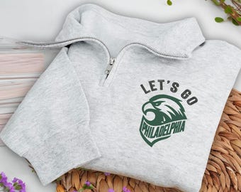 May include: A light gray sweatshirt with a quarter-zip collar. The sweatshirt features the text "LET'S GO PHILADELPHIA" with a green eagle logo. The garment is folded on a woven mat, with purple flowers and greenery in the background.