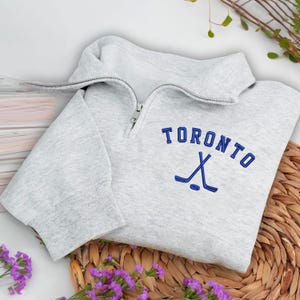 Op de afbeelding: Een lichtgrijs sweatshirt met een kraag met een kwartrits. Het woord "TORONTO" is blauw geborduurd, met gekruiste hockeysticks eronder. Het sweatshirt is op een geweven mat gevouwen, met paarse bloemen op de voorgrond.