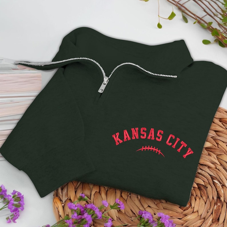 May include: Dark green quarter-zip sweatshirt with the words "KANSAS CITY" in red, arched over a red football graphic. The sweatshirt has a silver zipper and is displayed on a woven surface with purple flowers.