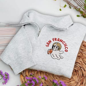 May include: A light gray sweatshirt with a quarter-zip neckline. The sweatshirt features an embroidered design of a ghost wearing a football helmet and holding a football, with the words "SAN FRANCISCO" in red.