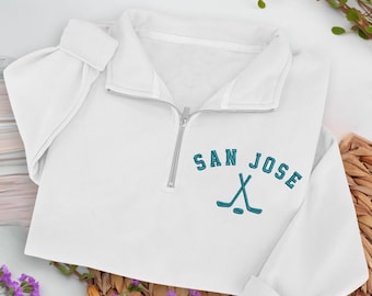 May include: White quarter-zip sweatshirt featuring the embroidered text "SAN JOSE" and two teal hockey sticks. The sweatshirt has a silver zip and folded cuffs. It is displayed on a woven surface with floral accents.
