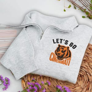 May include: A light gray sweatshirt with a quarter-zip collar. The front features the text "LET'S GO CINCINNATI" above an embroidered tiger graphic in black and orange. The sweatshirt is folded on a woven mat.