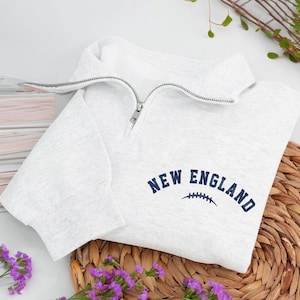 May include: A light gray sweatshirt with a quarter-zip collar. The words "NEW ENGLAND" are printed in navy blue, along with a football graphic. The sweatshirt is folded on a woven mat, with purple flowers in the foreground.