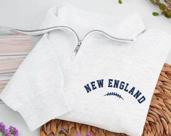 May include: A light gray sweatshirt with a quarter-zip collar. The words "NEW ENGLAND" are printed in navy blue, along with a football graphic. The sweatshirt is folded on a woven mat, with purple flowers in the foreground.