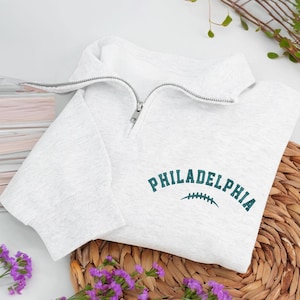 Może przedstawiać: Jasnoszara bluza z dekoltem zapinanym na zamek 1/4. Słowo "PHILADELPHIA" jest wyhaftowane w kolorze morskim, wygięte nad grafiką piłki nożnej w kolorze morskim. Bluza jest złożona na tkanej macie, z fioletowymi kwiatami na pierwszym planie.