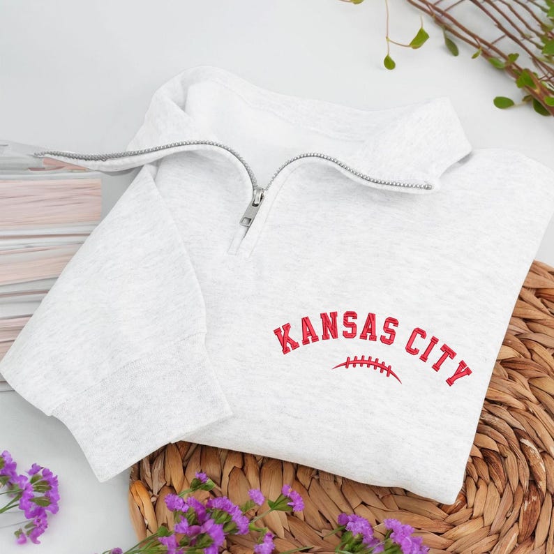 May include: A light gray sweatshirt with a quarter-zip neckline. The words "KANSAS CITY" are embroidered in red, with a red football graphic underneath. The sweatshirt is folded on a woven mat with purple flowers.