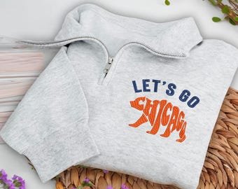 Może przedstawiać: Jasnoszara bluza z kapturem z kołnierzem zapinanym na zamek. Bluza ma napis "LET'S GO CHICAGO" w kolorze granatowym i pomarańczowym, z pomarańczową grafiką niedźwiedzia. Odzież jest złożona na tkanej powierzchni.