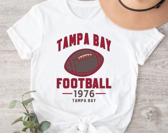 Pode incluir: Camiseta branca com o texto "TAMPA BAY FOOTBALL" e um gráfico de futebol americano. Abaixo do gráfico está "1976" e "TAMPA BAY". Um chapéu bege no fundo.