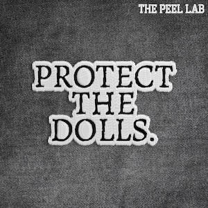 Peut inclure: Écusson brodé avec le texte "PROTECT THE DOLLS." en noir gras, bordé de blanc, sur un tissu gris. Le texte est superposé verticalement. Les mots "THE PEEL LAB" sont en haut.